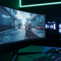 Egy ultra-széles gamer monitor közelről, amelyen egy gyors mozgású lövöldözős játék látható elmosódásmentesen, a háttérben sötét szobával és professzionális neon zöld-kék háttérvilágítással a falon.